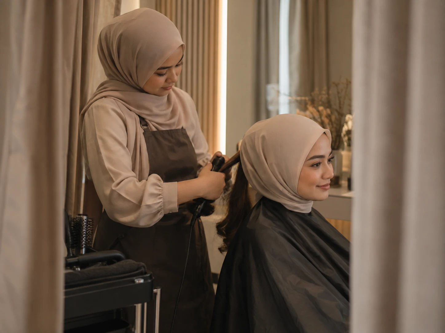 A female stylist styling a Muslimah client's hair in a private curtained area